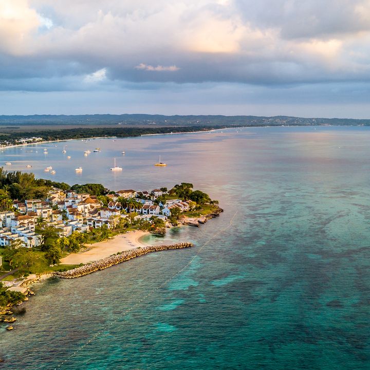 An aerial view over the coastline of Negril, Jamaica with buildings along the shore, boats dotting the water, and beautiful turquoise water.