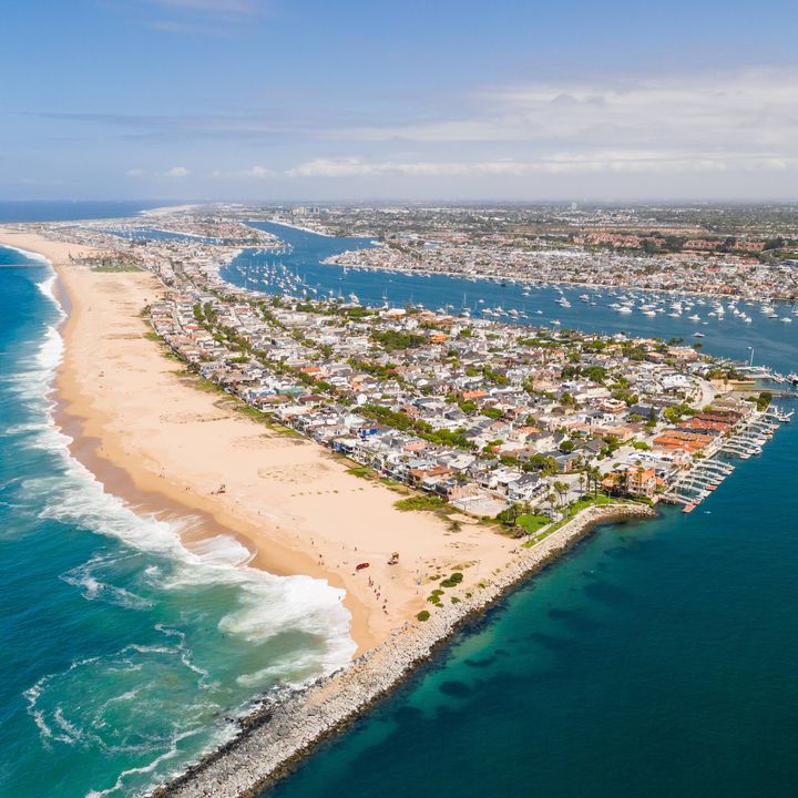 An aerial view over Newport Beach, including Balboa Island and the harbor, with a wide beach area and blue-turquoise water.