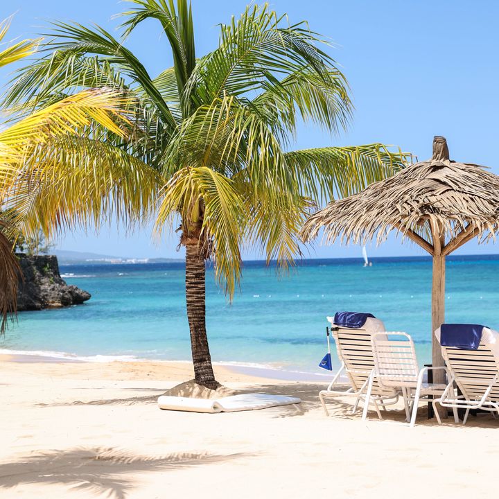 An empty tropical beach with white sand, swaying palm trees, blue water, and two empty lounge chairs.