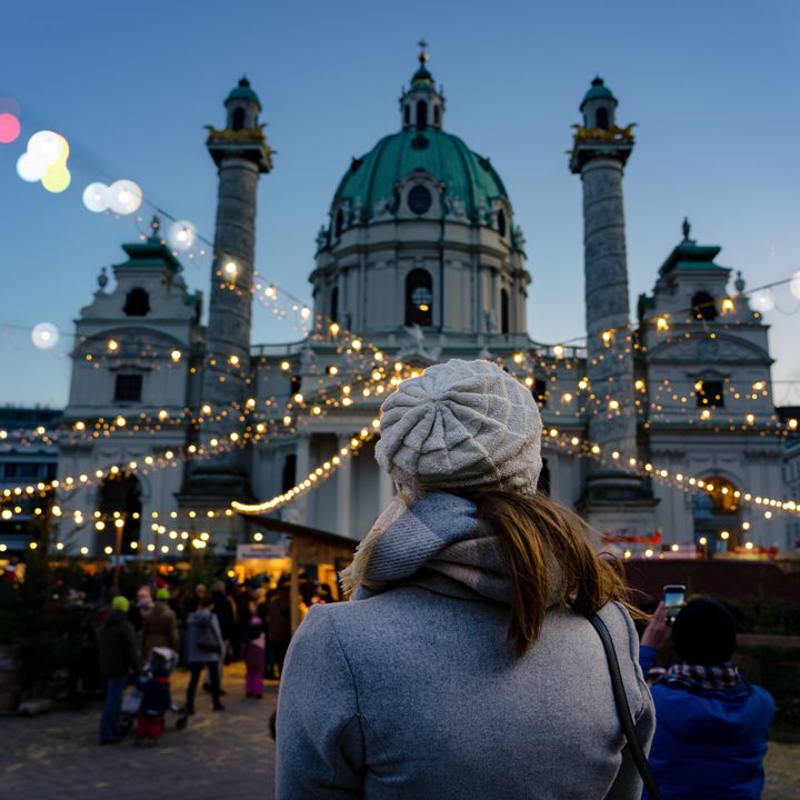 A woman is seen from behind, standing at the Ard Ardvent Christmas market in Vienna with string lights and booths in front of a church.