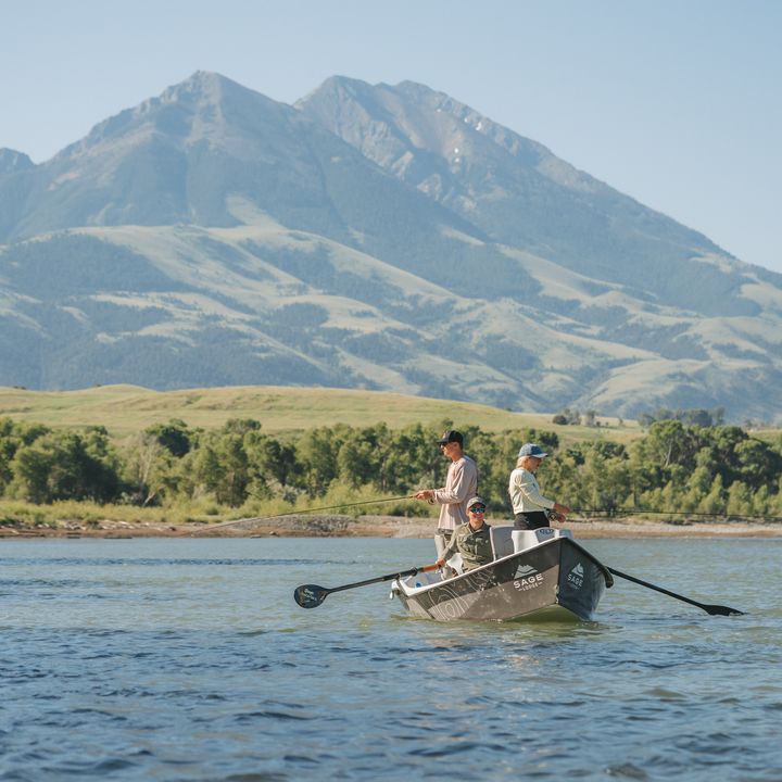 People fly fishing in a rowboat on the water in front of mountains in Montana.