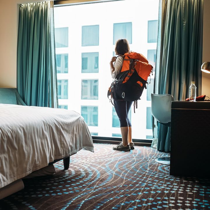 A woman stands at a window wearing a large backpack in a hotel room next to the bed.