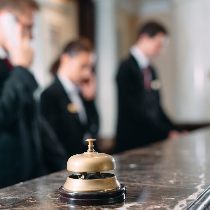 A marble hotel desk with a bell to ring for service on it, with people blurred out in the background on the phone.