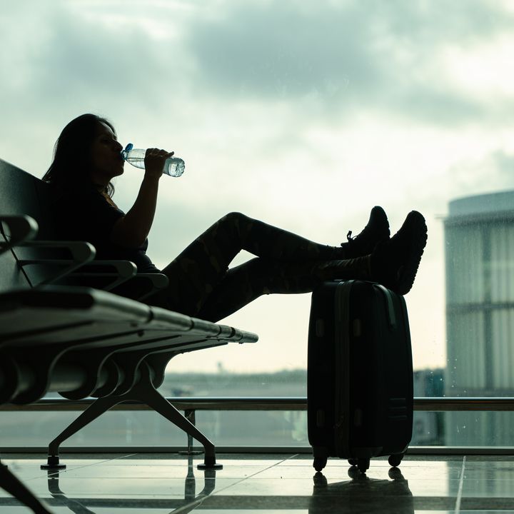 A low angle shot of a woman in dark light at the airport drinking a bottle of water while waiting for her flight with her feet propped up on her suitcase.