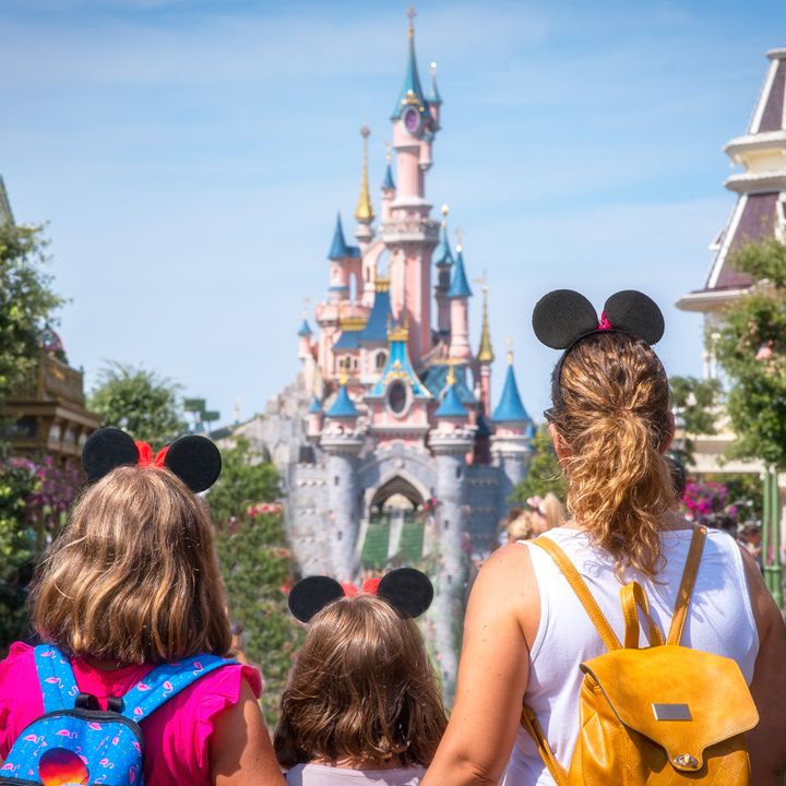 A woman and her two daughters stand in Disneyland looking at the castle, wearing backpacks.