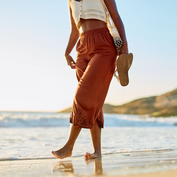 A woman is seen from the waist down walking on a tropical beach in the surf carrying her flip flop sandals.