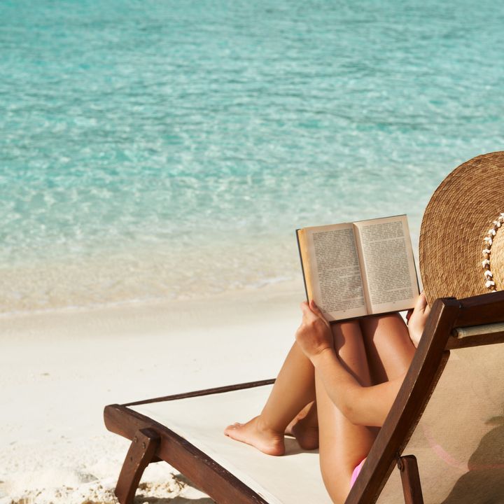 A woman is seen from behind reading a book while sitting on a beach chair facing crystal clean water and wearing a sun hat. 