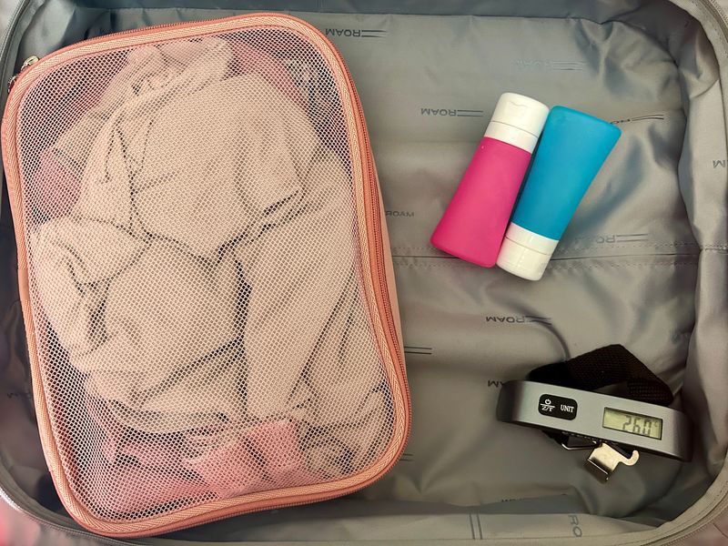 A suitcase half is filled with just a packing cube, blue and pink toiletry bottles, and a digital luggage scale.