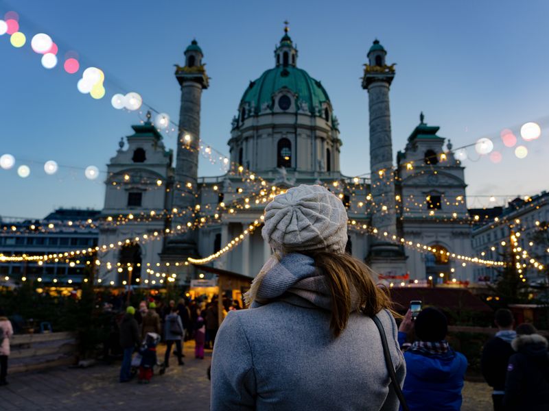 A woman is seen from behind, standing at the Ard Ardvent Christmas market in Vienna with string lights and booths in front of a church.