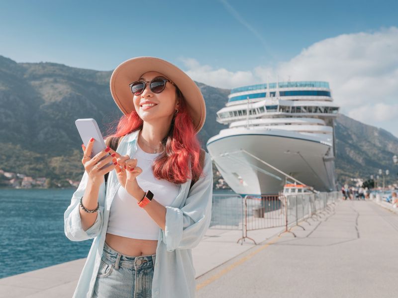A woman with pink hair wearing sunglasses and a hat on her smart phone and smiling next to a cruise ship.