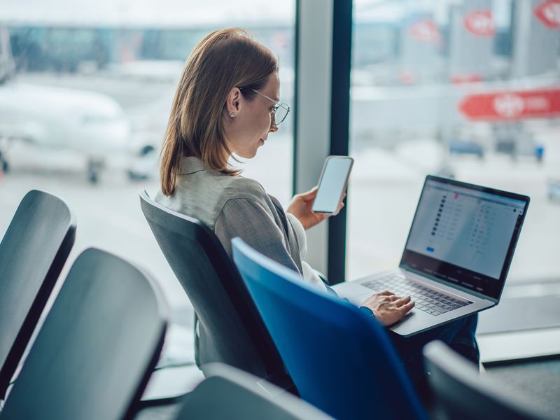 A woman sits in a chair at an airport holding her phone and looking at her laptop computer.