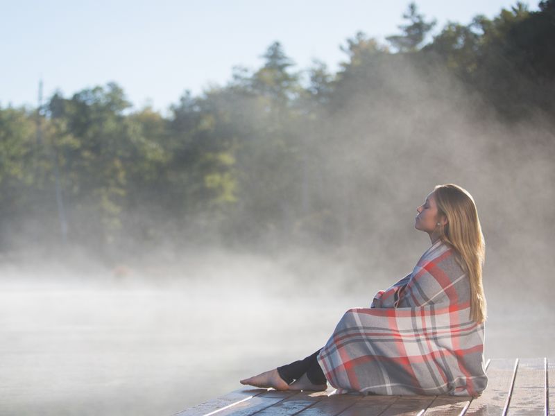 A woman sits on a dock at the water’s edge as steam rises off it, wrapped in a blanket.