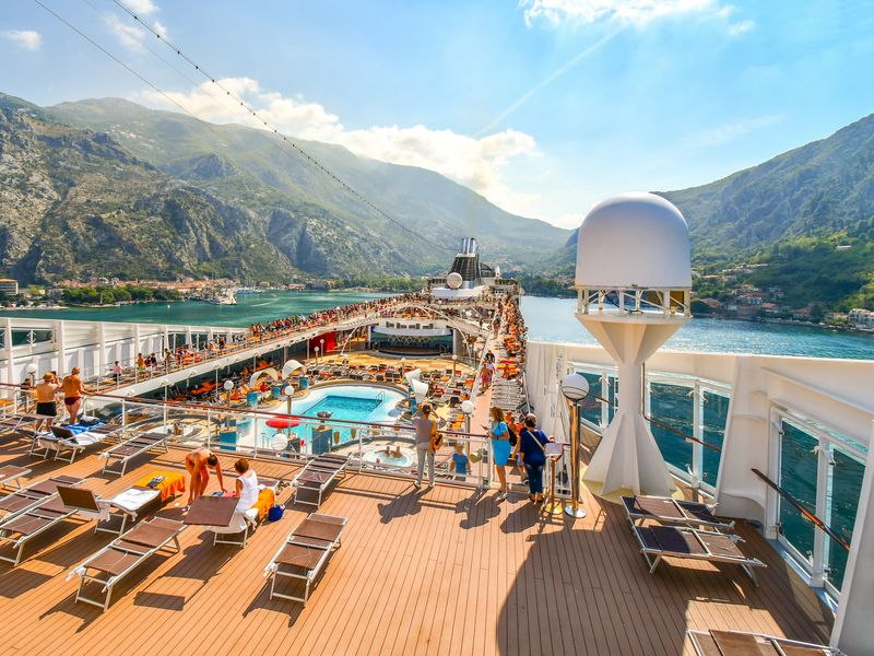 An aerial view over a top deck of a cruise ship with a pool and seating areas as the ship sails in a pretty Mediterranean setting.